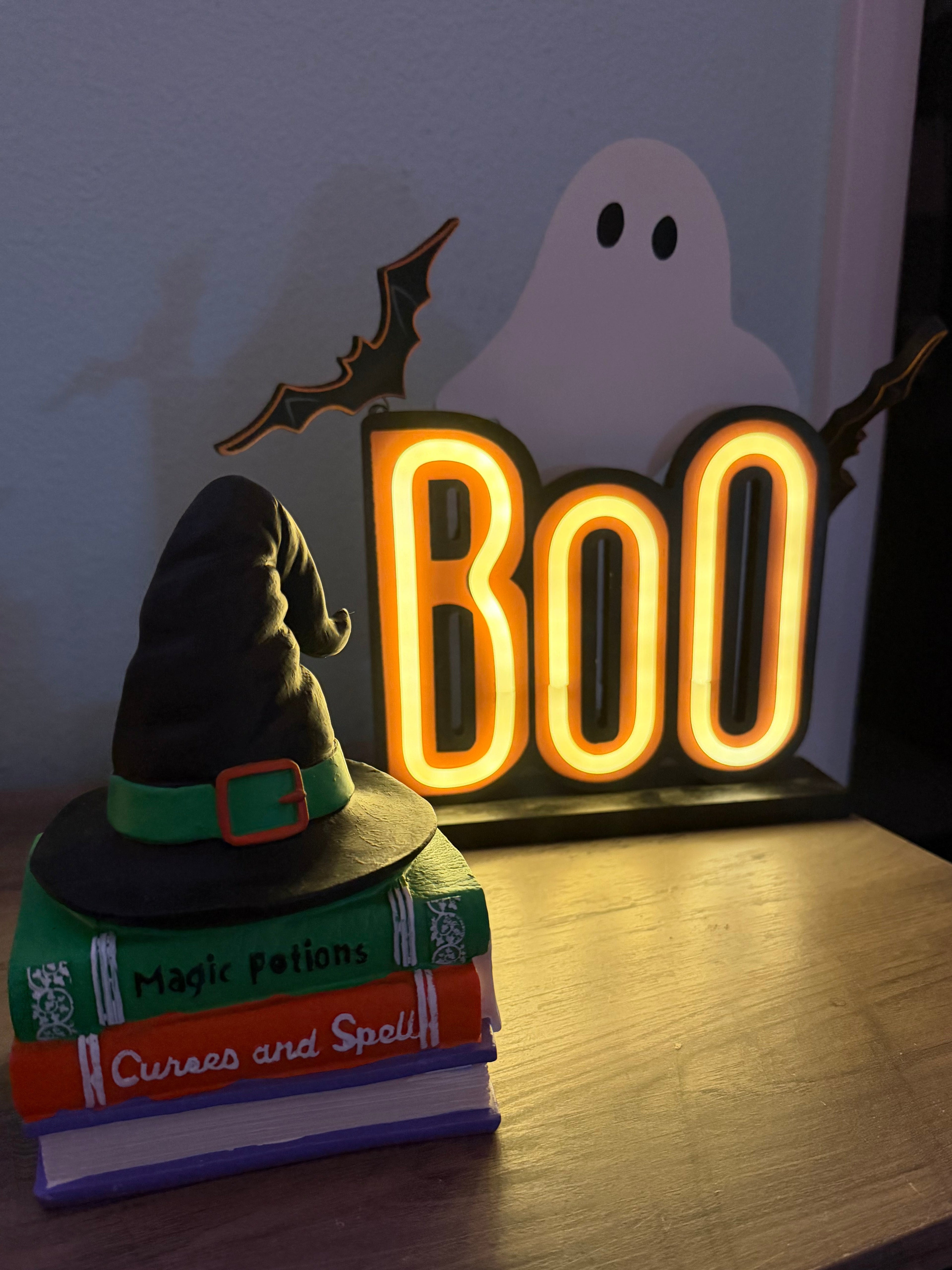 Neon 'BOO' sign with a ghost and bat on a white background, next to books and a green plush toy.