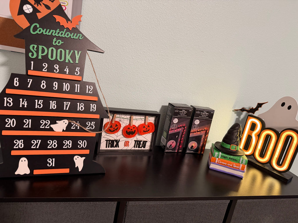 Halloween-themed decor items including a countdown calendar, framed picture, and neon sign on a table.